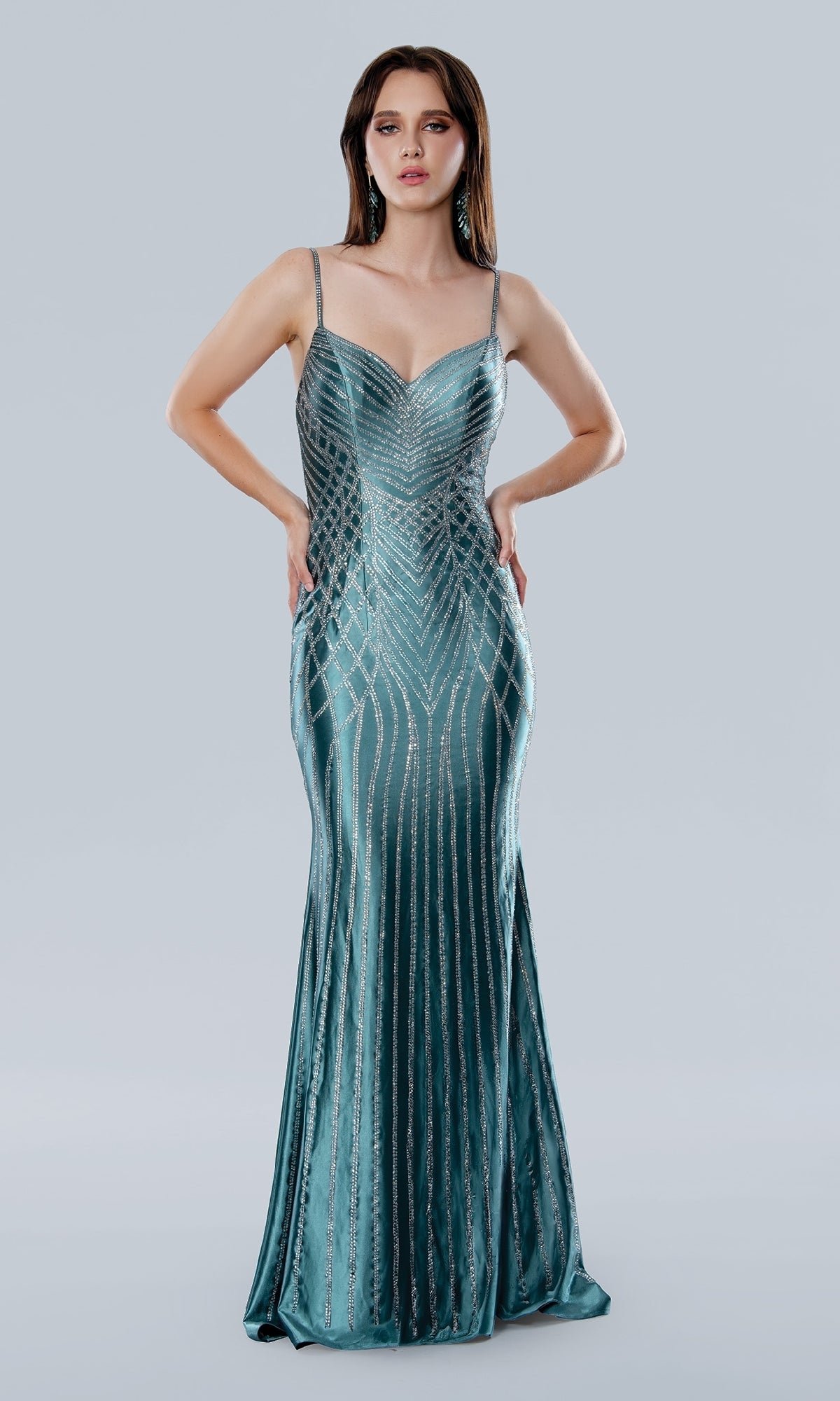 A woman poses in the Stella Couture 25185, a floor-length teal prom dress with silver geometric embellishments, against a plain light gray background.